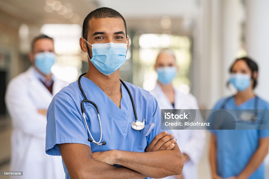 Team of doctors and nurses in hospital Confident multiethnic male nurse in front of his medical team looking at camera wearing face mask during covid-19 outbreak. Happy and proud indian young surgeon standing in front of his colleagues wearing surgical mask for prevention against coronavirus. Portrait of mixed race doctor with medical staff in background at hospital. Doctor Stock Photo