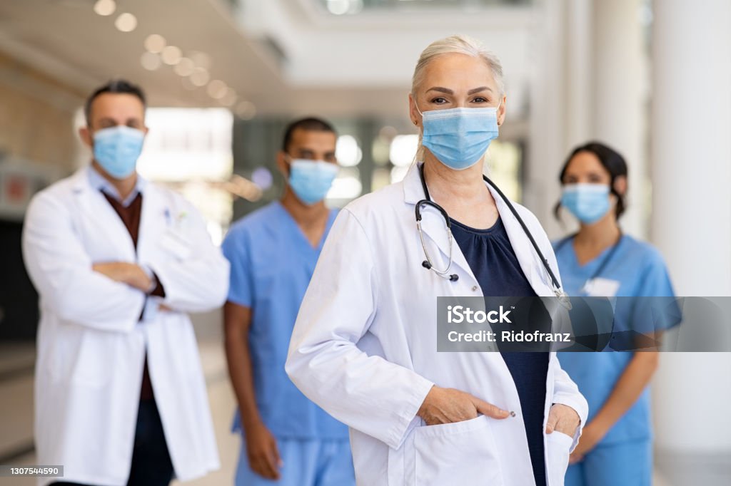 Satisfied doctor with medical staff looking at camera Portrait of smiling woman doctor standing in hospital with team in background wearing protective face mask during covid-19 pandemic. Female doctor in front of her medical staff with surgical mask at private clinic looking at camera. Healthcare workers team with general practitioner during corona virus pandemic. Doctor Stock Photo