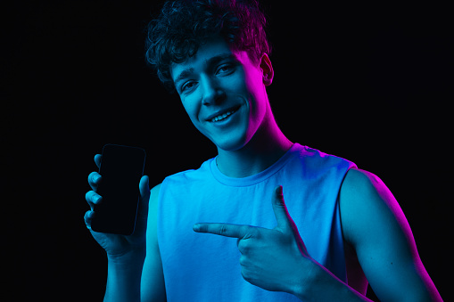 Pointing. Half-length portrait of young man with smartphone isolated on multicolored dark background in neon light. Concept of human emotions, facial expression, youth culture. Copy space for ad.