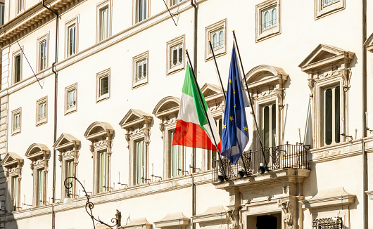 facade of Palazzo Chigi in Rome, seat of the Italian prime minister and government. The Italian and European flags on the balcony of the facade. Outdoors on a sunny day. Politics and democracy