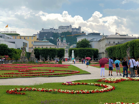 People in a green park with flowers and the popular historic castle on a hill in the background on a sunny day with a big cloud in the city of Salzburg in Austria June 10,2018