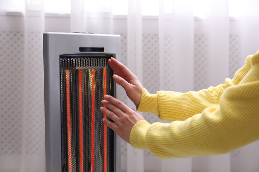 Woman warming hands near heater indoors, closeup