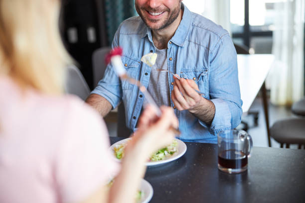Close up of bearded male person eating salad and smiling while sitting at the table and talking with lady