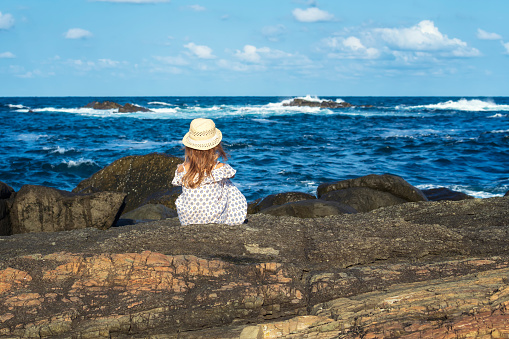 Rear view of a pretty little girl enjoys the sunny warm day at a rocky coastline