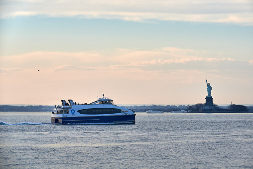 New York, NY - March 11 2021: In New York Harbor, a New York Waterway ferry passes in front of the Statue of Liberty in the late afternoon just before sunset.