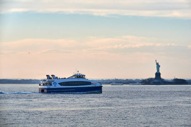 In New York Harbor, a New York Waterway ferry passes in front of the Statue of Liberty New York, NY - March 11 2021: In New York Harbor, a New York Waterway ferry passes in front of the Statue of Liberty in the late afternoon just before sunset. ferry stock pictures, royalty-free photos & images