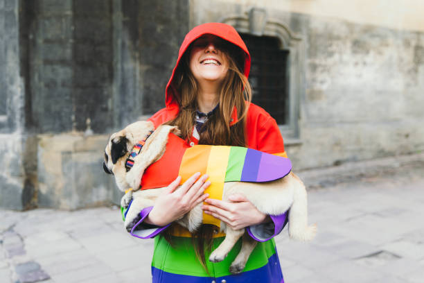 Happy young woman and her dog in rainbow coats walking in the sunny old city center enjoying the springtime Portrait of a woman with long hair holding her small cute dog - pug breed in multi colored rainbow raincoat enjoying the sunny spring day walking in the city centre enjoying the fresh blossom vibe in the air lgbtqcollection stock pictures, royalty-free photos & images