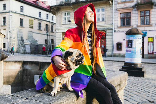 Woman with long hair and her small cute dog - pug breed in multi colored rainbow raincoat enjoying the sunny spring day walking in the city centre enjoying the fresh blossom vibe in the air