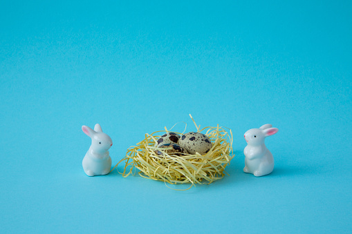 Figurines of two white Easter bunnies. Between them is a straw nest with quail eggs on a blue background. Space for inscriptions. Happy Easter!