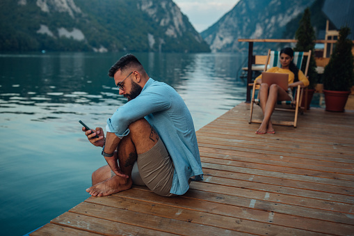 Man is sitting on pier and using phone while his girlfriend sitting on cozy chair behind him and using laptop