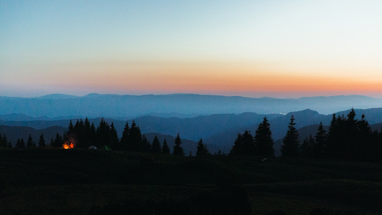 Panoramic view of the summer sunset above the blue mountain layered peaks, pine forest and the campsite with fireplace