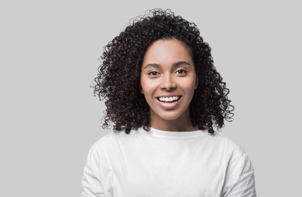 Mixed race woman wearing white t-shirt studio portrait Beautiful student girl with curly hair looking at camera. Studio shot. Isolated on gray background. People, beauty concept black hair stock pictures, royalty-free photos & images