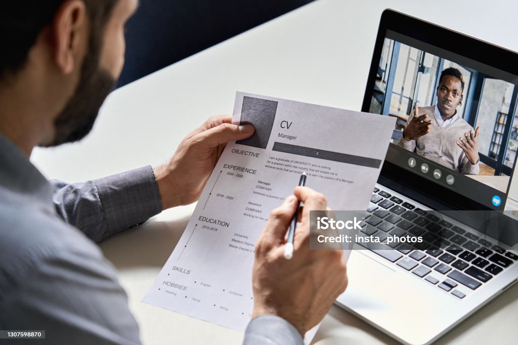 Male indian hr, recruiter or employer holding cv having online virtual job interview meeting with african candidate on video call. Distance remote recruitment conference chat. Over shoulder view. Recruitment Stock Photo