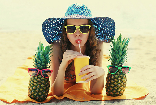 Portrait of young woman drinking a juice lying on a beach with funny pineapple wearing a straw hat