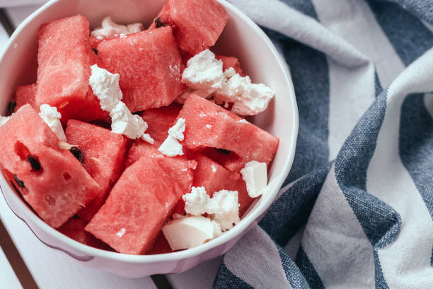 Delicious summer salad with slices of watermelon and feta cheese on white bowl with a striped towel on a wooden white background stock photo