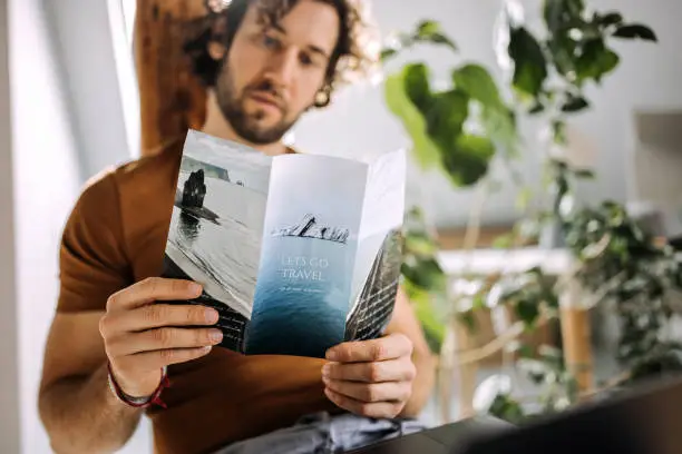 Photo of Young casually clothed man looking at travel flyer in his apartment