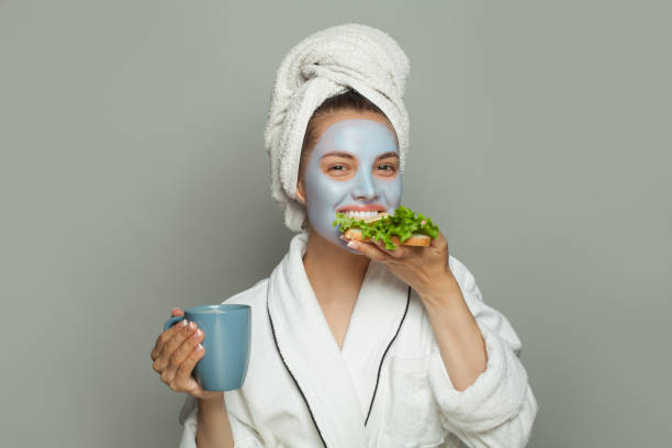bella donna in maschera cosmetica che mangia colazione e beve caffè. routine diurno femminile - bathrobe women cup towel foto e immagini stock