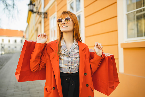 Portrait of redhead girl in modern coat with red shopping bags in the city