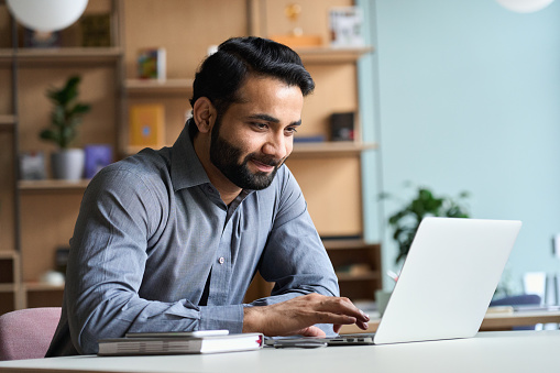 Un hombre de negocios indio sonriente trabajando en una computadora portátil en la oficina de casa. Joven estudiante indio o profesor remoto usando estudios remotos por computadora, capacitación virtual, viendo seminario web de educación en línea en la photo