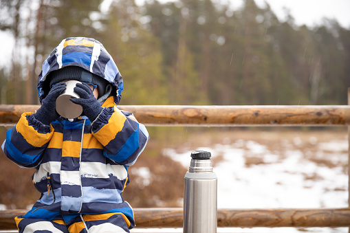 Young boy drinks hot beverage from a cup in a cold raini day. Resting, taking a break during hiking. Healthy living concept.