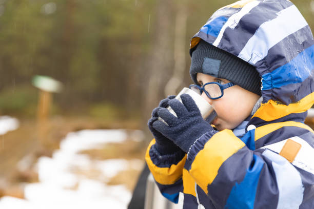 Young boy drinks hot beverage from a cup in a cold raini day. Resting, taking a break during hiking. Healthy living concept.