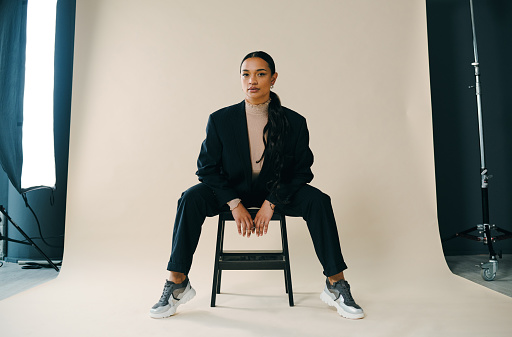 Studio portrait of a beautiful young woman sitting on a chair during a photoshoot against a brown background