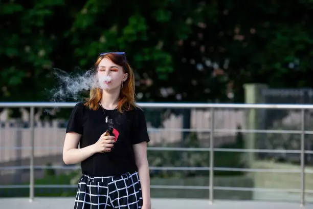 Vape teenager. Young pretty white caucasian girl with glasses on her head an electronic cigarette on the street in the summer. Deadly bad habit.