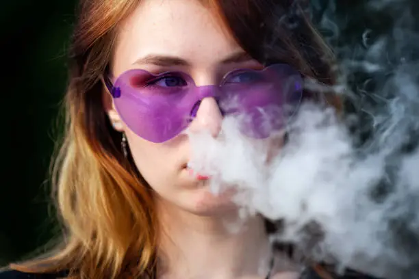Vape teenager. Young pretty white caucasian girl in purple glasses on her head an electronic cigarette on the street in the summer. Deadly bad habit.