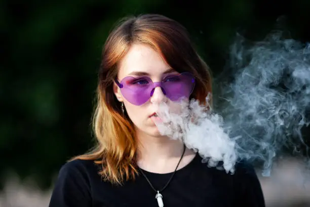 Vape teenager. Young pretty white caucasian girl in purple glasses on her head an electronic cigarette on the street in the summer. Deadly bad habit.