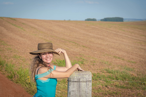 Brazilian woman wearing a hat propped up on a rural property fence. Lifestyle. Girl posing for photo in farm area in Brazil. Rural landscape.