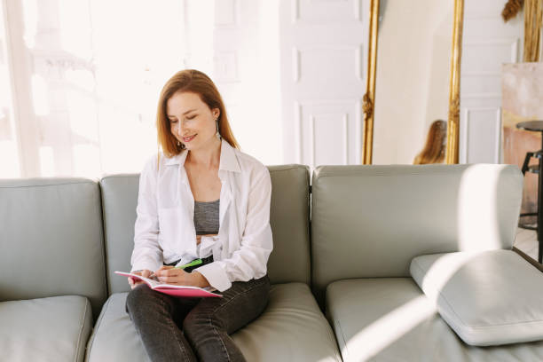 istock A pretty woman sits on a sofa at home and writes in a notebook on her lap 1307634700