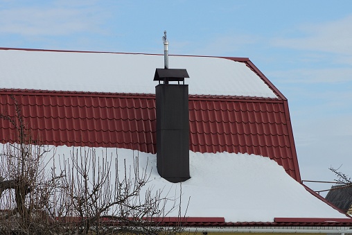 brown metal chimney on a red tiled roof of a private house under white snow against a blue sky on a winter street