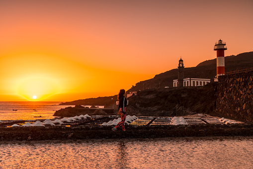 A young woman walking in the orange sunset in the salina and in the background the Fuencaliente Lighthouse on the route of the volcanoes south of the island of La Palma, Canary Islands, Spain