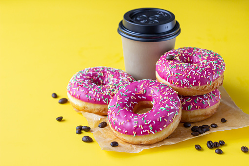 Takeaway coffee cup with coffee beans and pink glaze donuts with colorful sprinkles, pink background. Selective focus.