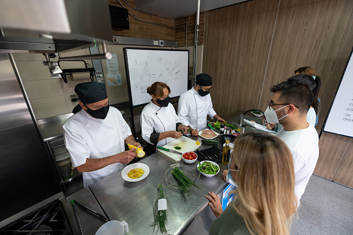 Group of students and chefs wearing facemasks in a cooking class during the COVID-19 pandemic - education concepts