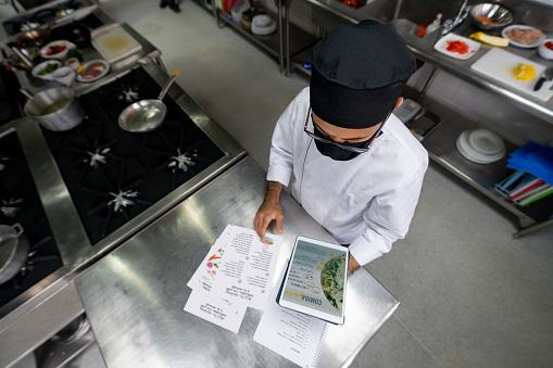 Chef wearing a facemask and designing a menu after reopening his restaurant during the COVID-19 pandemic. **DESIGN ON SCREEN AND DOCUMENTS WERE MADE FROM SCRATCH BY US**