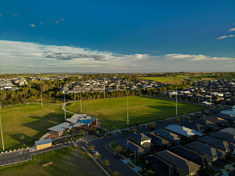 High angled photo of green grassed suburban sporting fields surrounded by housing estate on sunny afternoon, sun setting over mountains casting shadows.