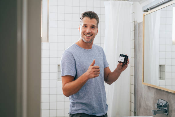 Portrait of a handsome happy young Caucasian man giving thumbs up and looking at camera while holding a face cream jar after a morning shave.