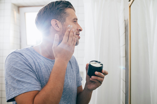 Handsome happy young Caucasian man applying face cream after a morning shave.