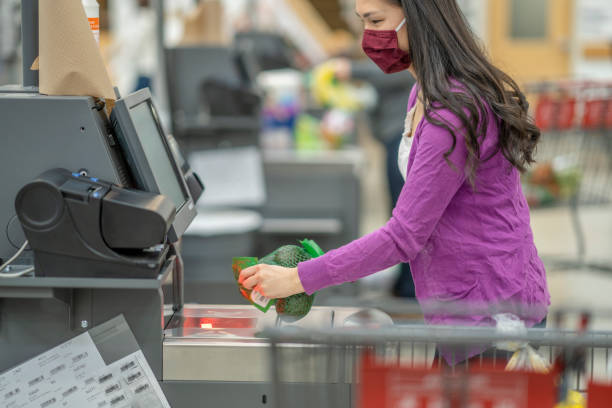 Female Adult at the Grocery Store Self Check Out Service A young female adult scanning a bag of avocados at the grocery store self check out service. She is wearing a face mask during the coronavirus pandemic to prevent the spread of germs. self checkout photos stock pictures, royalty-free photos & images