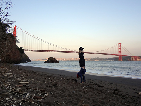 Man wearing hat, hoodie, long pants and shoes Handstands on beach in front of the Golden Gate Bridge on the Marin side with San Francisco, California in the distance.