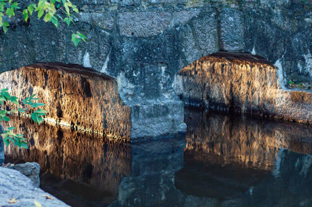 Old, small stone bridge close-up across small river, warm sunlight reflected from arches bridge, calm water surface.