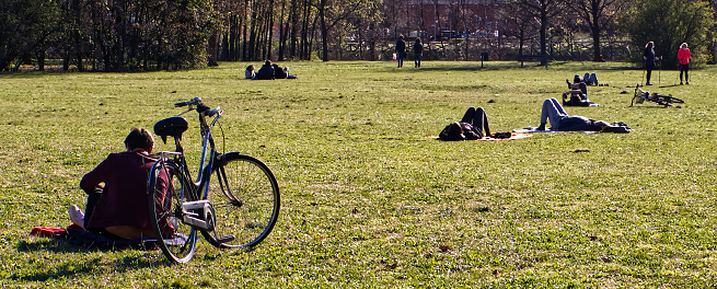 Bologna - Italy - March 17, 2021: People relaxing on the park lawn on a sunny day during the covid-19 pandemic. Living with Covid-19. Resting in the park maintaining social distance and medical masks during the coronavirus pandemic.