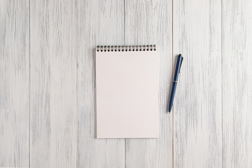 An overhead view of a mockup of a blank notepad and an automatic ballpoint pen on a light painted wooden surface. Selective focus. Copy space.