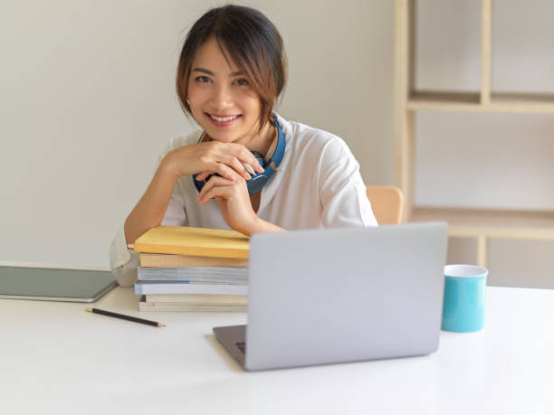 istock Female student smiling to camera while sitting at study table wit laptop and stack of books 1307705633
