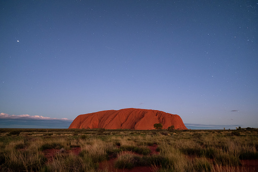 Northern Territory, Australia - March 29, 2016: Night has fallen in the heart of the Australian Outback, and countless stars (and possibly a shooting star) gently illuminates the massive form of Uluru that looms majestically on the horizon.