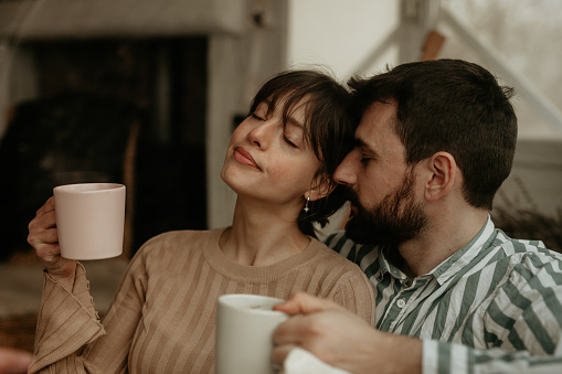 Beautiful young adult couple sitting in their living room, next to the window, enjoying their coffee and tea while cuddling, having their eyes closed and a gentle smile on their face.