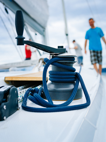 Winch of main sail with rope and handle on sailboat. Sailing team in background. Model released.
