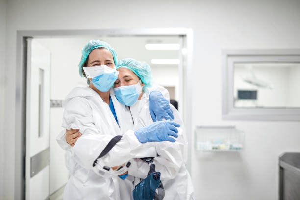 Healthcare coworkers embracing each other in ICU Healthcare coworkers embracing each other in ICU. Doctors are wearing protective white coveralls. They are standing outside hospital ward. frontline worker mask stock pictures, royalty-free photos & images