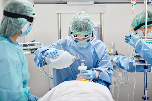 Medical professionals intubating female patient in ICU. Frontline workers wearing blue protective coveralls. They are at hospital during pandemic.